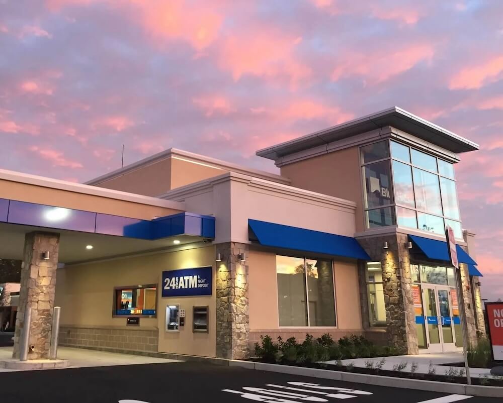 The image captures a modern bank branch exterior at twilight with a stunning pink and orange cloud-filled sky in the background. The building features a contemporary design with a mixture of stone columns and neutral tones, complemented by blue awnings over the windows and doors. Visible signs highlight the availability of 24-hour ATM service and night deposits, indicating the bank’s emphasis on convenience and accessibility for its customers. The overall atmosphere is welcoming, designed to attract customers with its aesthetic appeal and the practical services it offers.