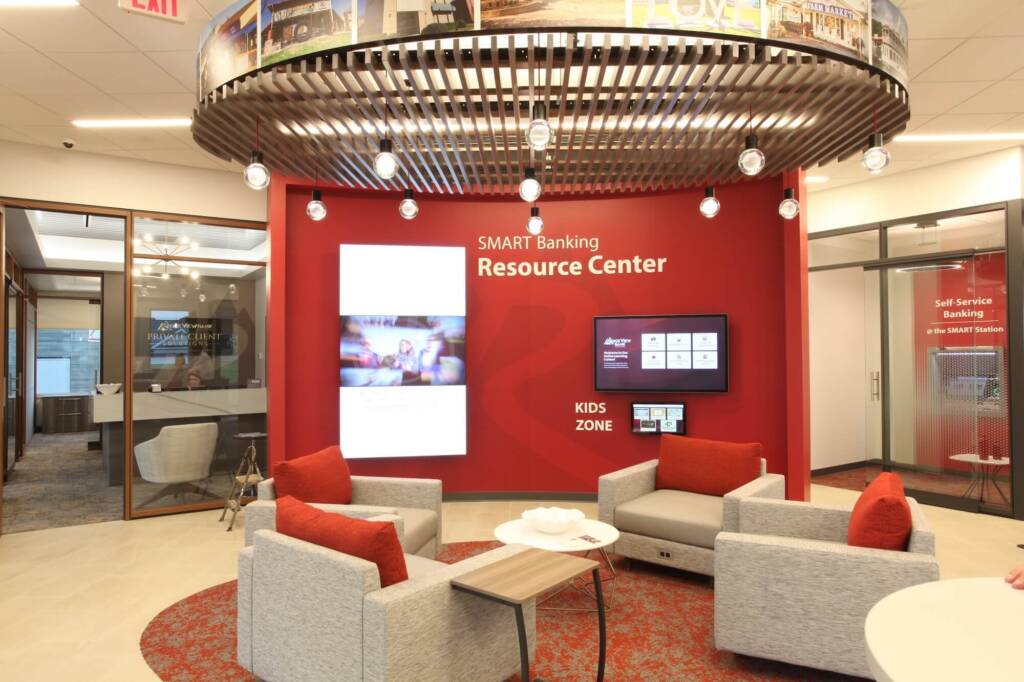 Bank interior seating area with gray chairs, a round table, and a circular overhead light fixture. Digital screens, a red accent wall, and glass-walled offices are visible in the background.