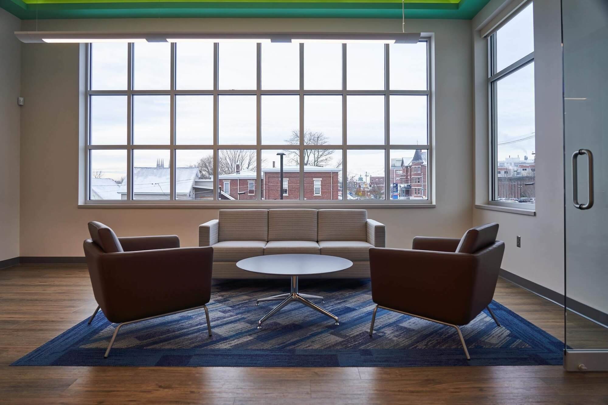 Spacious lounge area with large windows, modern brown chairs, a gray couch, and a round coffee table. The room is filled with natural light, reflecting inviting credit union and bank interior design.