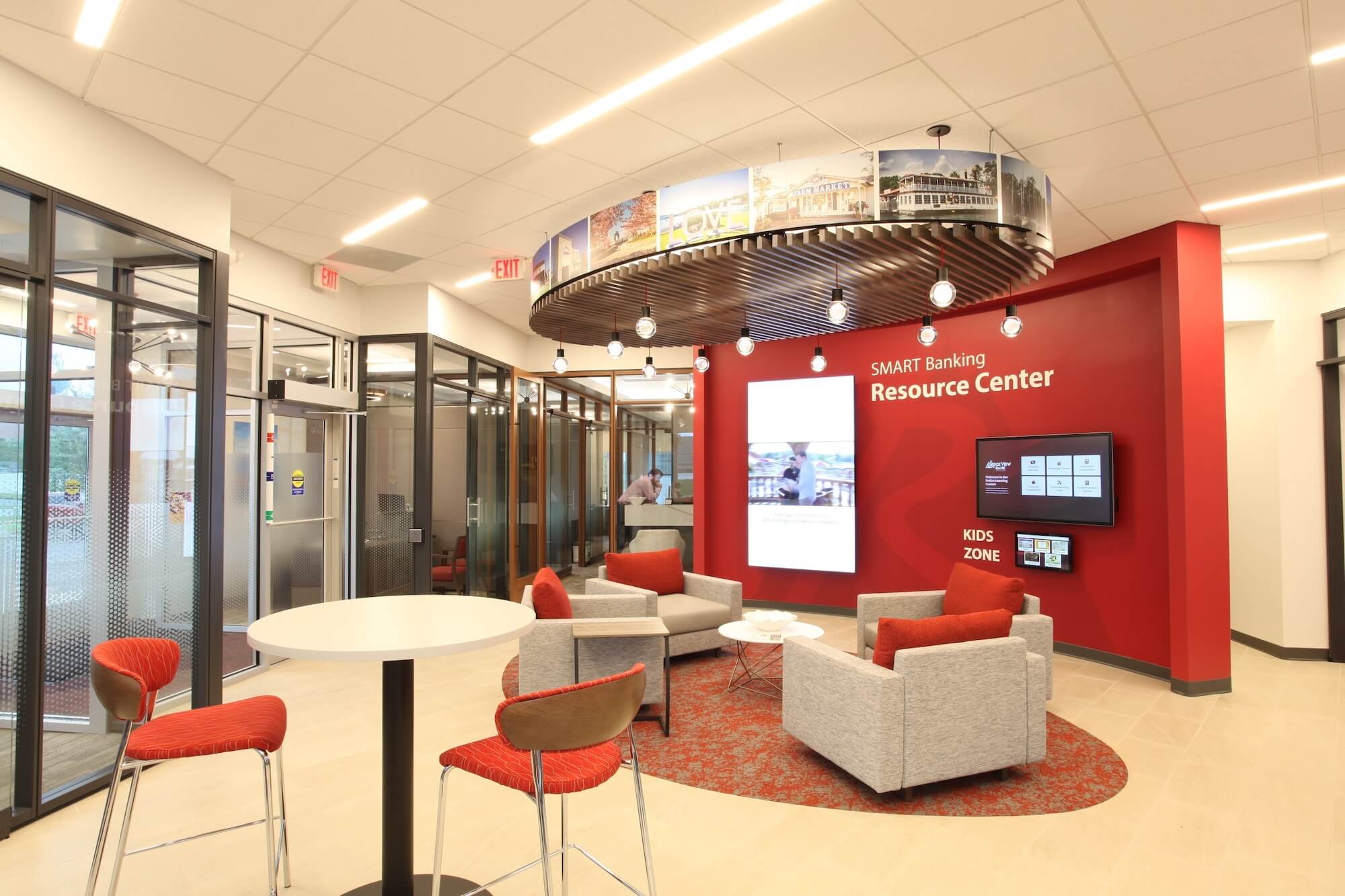 Modern bank interior with a red accent wall, gray chairs with red pillows, a white table, pendant lights, and a round table with two red chairs.